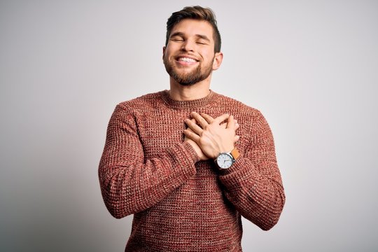 Young blond man with beard and blue eyes wearing casual sweater over white background smiling with hands on chest with closed eyes and grateful gesture on face. Health concept.
