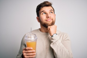 Fototapeta premium Young blond man with beard and blue eyes drinking healthy orange smoothie serious face thinking about question, very confused idea