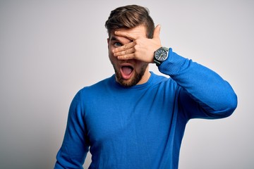 Young handsome blond man with beard and blue eyes wearing casual sweater peeking in shock covering face and eyes with hand, looking through fingers with embarrassed expression.