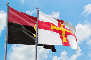 Guernsey and Angola flags waving in the wind against white cloudy blue sky together. Diplomacy concept, international relations.