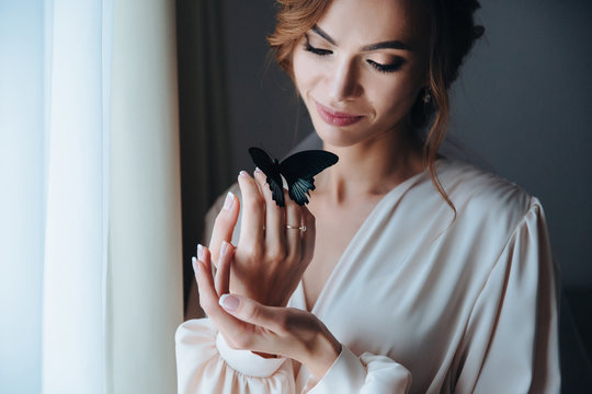 Close up portrait of a beautiful gentle bride in a beige peignoir with a butterfly with black and blue wings sits on the hand of the bride