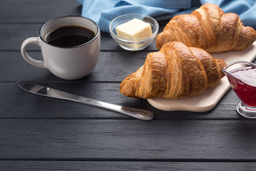 Fresh croissants on a black wooden table. Served with coffee, coffeepot, butter, jam, blue napkin. Fresh French Baked Croissants. Warm Fresh Buttery Rolls. Free space for text. 