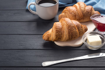 Fresh croissants on a black wooden table. Served with coffee, coffeepot, butter, jam, blue napkin. Fresh French Baked Croissants. Warm Fresh Buttery Rolls. Free space for text. 