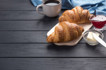 Fresh croissants on a black wooden table. Served with coffee, coffeepot, butter, jam, blue napkin. Fresh French Baked Croissants. Warm Fresh Buttery Rolls. Free space for text. 