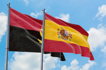 Spain and Angola flags waving in the wind against white cloudy blue sky together. Diplomacy concept, international relations.