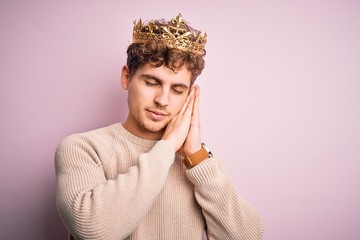 Fototapeta na wymiar Young blond man with curly hair wearing golden crown of king over pink background sleeping tired dreaming and posing with hands together while smiling with closed eyes.