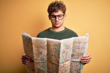 Young blond tourist man on vacation with curly hair holding city map over yellow background with a confident expression on smart face thinking serious