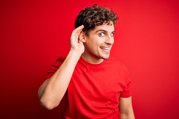 Fototapeta na wymiar Young blond handsome man with curly hair wearing casual t-shirt over red background smiling with hand over ear listening an hearing to rumor or gossip. Deafness concept.