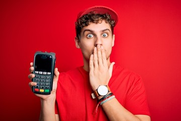 Young blond man with curly hair wearing cap holding dataphone over red background cover mouth with...