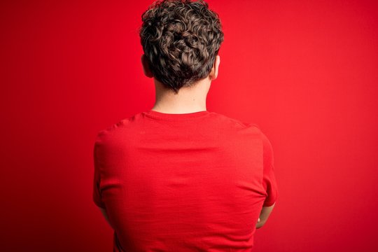 Young blond handsome man with curly hair wearing casual t-shirt over red background standing backwards looking away with crossed arms