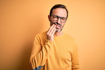 Middle age hoary man wearing casual sweater and glasses over isolated yellow background touching mouth with hand with painful expression because of toothache or dental illness on teeth. Dentist