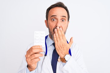 Middle age doctor man holding pills standing over isolated white background cover mouth with hand shocked with shame for mistake, expression of fear, scared in silence, secret concept