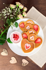 Homemade heart shaped pancakes with strawberry cream and berry slices on a white plate on a wooden table. Rustic style breakfast or brunch for Valentine's Day. Top view, vertical orientation
