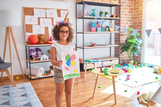 Beautiful toddler wearing glasses and unicorn diadem standing holding draw smiling at kindergarten