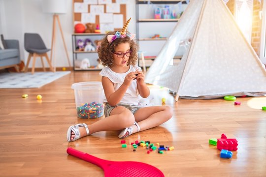 Beautiful toddler wearing glasses and unicorn diadem sitting on the floor playing with building blocks at kindergarten