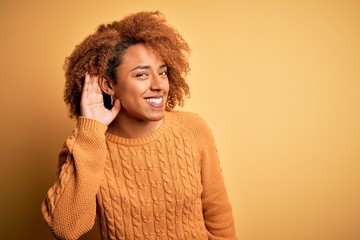 Young beautiful African American afro woman with curly hair wearing casual sweater smiling with hand over ear listening an hearing to rumor or gossip. Deafness concept.