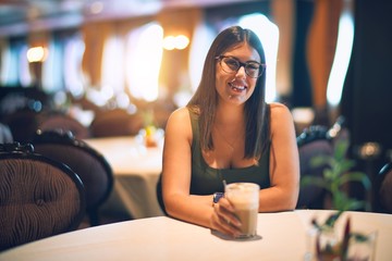 Young beautiful woman smiling happy and confident. Sitting with smile on face holding glass of coffee at restaurant