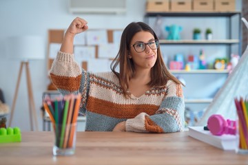 Young beautiful teacher woman wearing sweater and glasses sitting on desk at kindergarten Strong person showing arm muscle, confident and proud of power