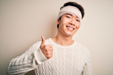 Young handsome chinese man injured for accident wearing bandage and strips on head doing happy thumbs up gesture with hand. Approving expression looking at the camera showing success.