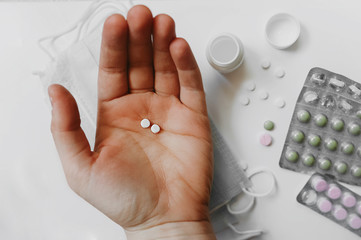 in the light-skinned hand of a man, two small white tablets against a background of blurred medical items, colored tablets in the package and respiratory masks