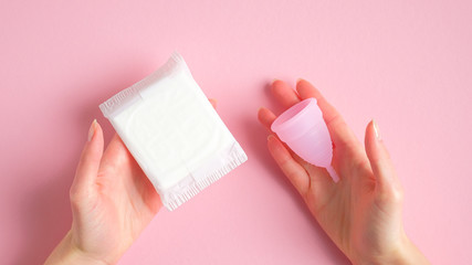 Female hands holding sanitary pad and menstrual cup over pink background. Different types of menstrual hygiene products comparison. Critical days, menstruation cycle, female healthcare concept