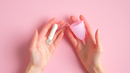 Female hands holding tampon and menstrual cup over pink background. Different types of menstrual hygiene products comparison. Critical days, menstruation cycle, female healthcare concept - obrazy, fototapety, plakaty