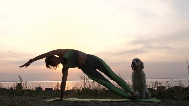 Sporty slim female doing side plank while exercising yoga on sea shore at sunset, golden retriever pet sitting next helping the workout. Mature woman practicing yoga in nature with dog partner