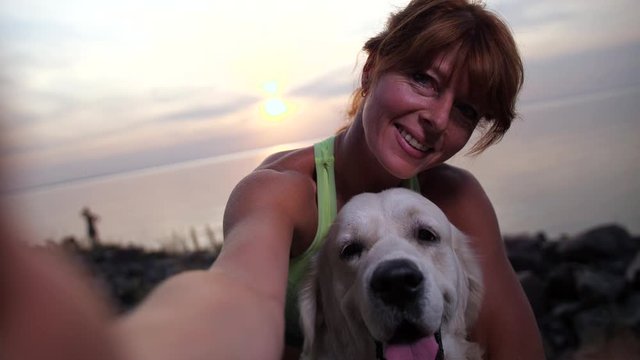 Closeup portrait of attractive sporty redhead female posing for selfie photo on smartphone together with cute golden retriever at sea sunset. Pet owner and dog photographing on sea shore after workout