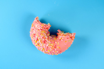 Pink frosted donut with colorful sprinkles with bite missing. On blue background .