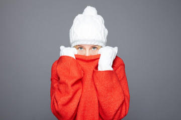 How chilly. Puzzled blue eyed lady hugs herself, trembles from cold, walks on windy weather, shakes and clenches teeth, dressed in red sweater, stands without coat, isolated on grey studio wall.