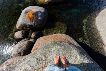 Point of view looking down at mans feet on edge of lichen covered granite rock overhanging the sea 