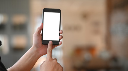 Man's hand holding crop black smartphone with white blank screen and pointing finger on button with modern living room as blurred background.