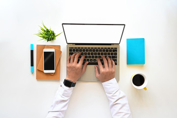 Top view of creative man in white shirt typing on white blank screen laptop on the modern working desk. Smartphone with black empty display, coffee cup, notebook, diary and potted plant putting on it.