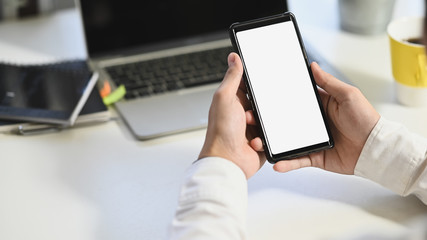 Cropped shot of business man while using/holding white blank screen smartphone with laptop, coffee cup and notebook putting on white table as background.