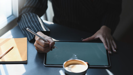 Cropped shot of young executive woman's hands holding a pen while drawing business plan by computer tablet at the modern working desk with comfortable office as background.