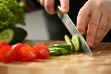 Cook holds knife in hand and cuts on cutting board green cucumber for salad or fresh vegetable soup with vitamins. Raw food and vegetarian recipe book in modern society popular concept.