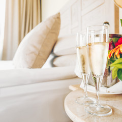 Two glasses of champagne in the upscale hotel room. Dating, romance, honeymoon, valentine, getaway concepts. Square. Light warm toning