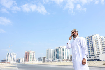 Fototapeta premium Middle east business mand wearing white dress talking on smart phone standing on street over buildings background, Bahrain.