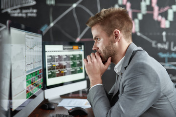 Fototapeta na wymiar Keep growing with smart ideas. Portrait of successful young trader looking focused while sitting in front of multiple monitors in the office. Blackboard full of chart and data analyses in background.
