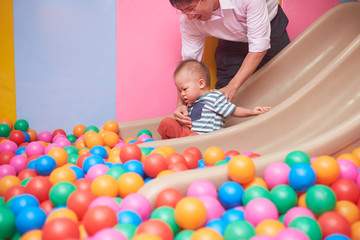 Cute little Asian toddler boy playing on a slide in ball pit full of colorful plastic ball, Dad and son having fun in children play center, Businessman father playing with son after working time