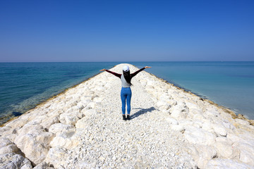 Woman standing on the pier white rocks showing hands up to blue sky over sea water background, Bahrain.