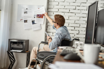 Working day. Young busy male office worker in a wheelchair putting some documents on white board while sitting near printer at his workplace in the bright modern office