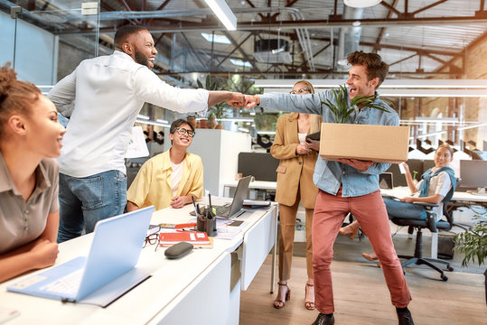 Welcome to our new office. Young handsome man in casual wear giving fist bump to his new coworker while standing in the modern office with other colleagues