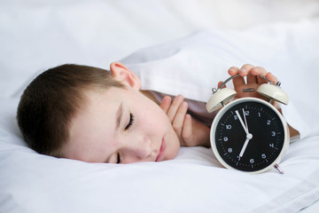 The boy is sleeping and holding an alarm clock with his hand. A baby in a white bed sleeps in the morning.