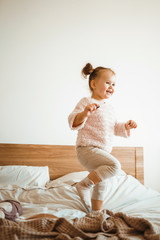 Fototapeta premium Happy little girl having fun in bed while being at home. Cute baby playing and jumping baby in the bedroom. Happy loving family.