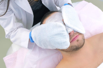 Fototapeta premium Cosmetologist wipes white moisturizing mask from man's face using terry mittens in beauty clinic, face closeup. Beauty industry concept. Guy is lying on couch. Beautician making procedure.
