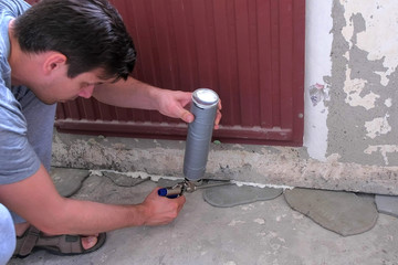 Young man worker fills crack between wall and cement floor with mounting foam in room under the battery. Repair and construction work of the house. Rough finish of the room.