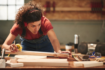 Young concentrated curly woman in ear defenders and overall measuring wooden plank with tape measure - obrazy, fototapety, plakaty