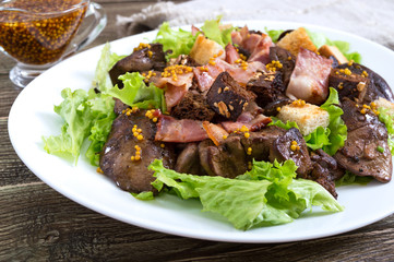 Warm healthy salad of chicken liver, rye croutons, smoked bacon, green salad and mustard sauce in a white plate on a wooden background. Close up