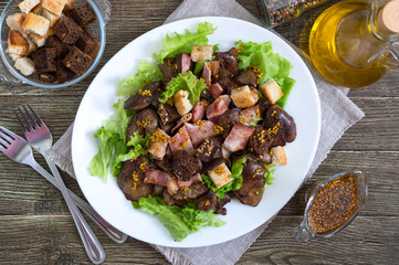 Warm healthy salad of chicken liver, rye croutons, smoked bacon, green salad and mustard sauce in a white plate on a wooden background. Top view.